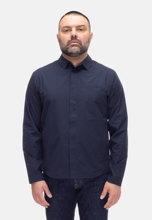 Uomo con capelli corti scuri e barba che indossa una camicia blu scura abbottonata e jeans scuri, in piedi davanti a uno sfondo chiaro e uniforme.