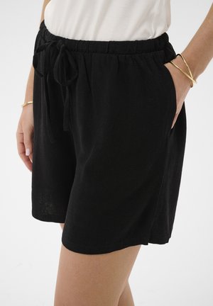 Kvinde iført sorte elastiske shorts med snøre i taljen, hvid top og gyldne armbånd på begge håndled, hænderne nær lommerne.