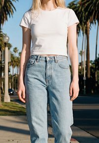 Jeune personne portant un t-shirt court blanc et un jean bleu clair taille haute, debout en extérieur avec des palmiers et des lampadaires en arrière-plan.