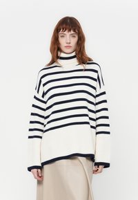 Maglione a collo alto a righe in crema e blu navy. Presenta una vestibilità rilassata, una texture a coste e maniche larghe, con un orlo leggermente asimmetrico.