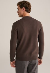 Göndör barna hajú férfi, aki háttal áll, sötétbarna pulóvert és szürke kockás nadrágot visel egy semleges háttér előtt.