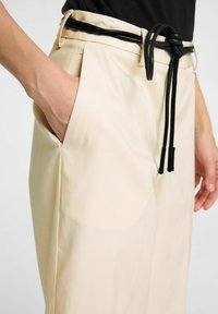 Beige vida byxor med slät yta, utrustade med ett svart knytbälte, sidfickor och veckad front för extra detalj.