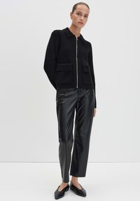 Vrouw in een zwarte ritsgebreide trui met voorzakken, zwarte leren rechte broek en zwarte puntige flats, staand tegen een witte achtergrond.