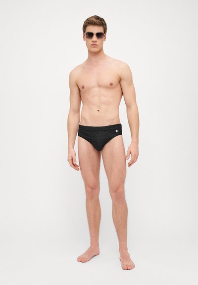 Caleçons de bain noirs avec un motif texturé, dotés d'une ceinture douce et d'un petit logo sur le côté, modèle debout pieds nus.