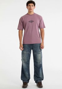 Kurzärmeliges, lavendelfarbenes T-Shirt mit dem "ORIGINALS"-Logo, kombiniert mit lockeren, ausgewaschenen blauen Cargo-Hosen mit mehreren Taschen und gesteppten Details.