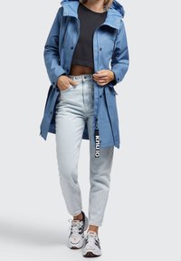 Blå vattentät jacka med huva, tryckknappar och dragsko, kombinerad med ljusa jeans och sportiga sneakers. Texturen ser slät ut.