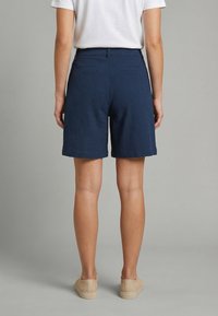 Persona con pantalones cortos de textura azul marino, camisa blanca de manga corta y zapatos beige sin cordones, de pie contra un fondo gris liso.