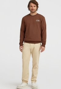 Man in een bruin O'Neill sweatshirt, beige broek en witte sneakers, staande tegen een effen witte achtergrond.