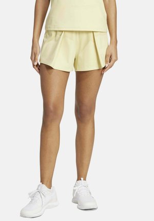 Femme portant un short plissé jaune pâle et un haut assorti, associés à des baskets de sport blanches, debout sur un fond uni.