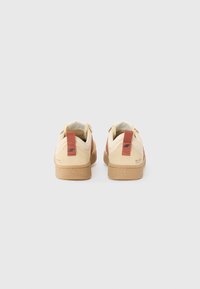 sneakers de camurça bege com uma sola texturada em tom de caramelo, apresentando uma aba vermelha na parte de trás e um logótipo. O design é minimalista e de cano baixo.