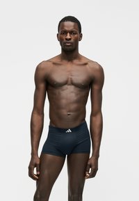 Sportisks tumšādains vīrietis stāv pretī skatītājam, valkājot melnas Adidas bokseršortus uz vienkārša baltā fona.