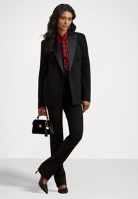 Melna divrindu žakete ar satīna lapiņām, sarkana rakstota blūze, melni bikses un melna rokassoma ar zelta akcentiem uz viendabīga fona.