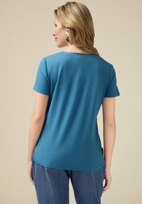 T-shirt teal cu mânecă scurtă dintr-un material moale, cu guler rotund și tiv curbat. Asortat cu blugi decolorati, văzut din spate.