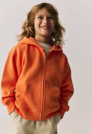 Kind mit schulterlangem Haar lächelnd, trägt eine orangefarbene Kapuzenjacke mit Reißverschluss und helle Hose, Hände in den Taschen, vor einem einfarbigen Hintergrund.