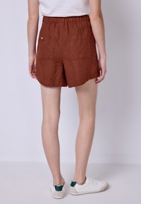 Shorts in lino marrone con elastico in vita, dotati di due tasche posteriori e un fit comodo. Abbinati a sneakers bianche.