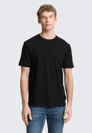 Junger Mann mit lockigem Haar, der ein schlichtes schwarzes kurzärmeliges T-Shirt und blaue Jeans trägt, steht vor einem hellgrauen Hintergrund.