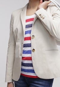 Personne portant un blazer beige par-dessus une chemise rayée rouge, blanc, bleu et gris, associée à un jean bleu, tenant un sac blanc sur l'épaule.