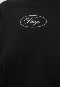 Svart stoff med hvitt ovalt logo som inneholder kursiv tekst "Hugo" sentrert nær toppen.