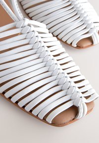 Sandales en cuir blanc avec un design tressé, bout ouvert et semelle plate. La texture est lisse avec une doublure intérieure beige visible.