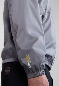 Grau Windbreaker mit elastischem Saum, Reißverschlusstasche und gummiertem Logo. Hergestellt aus strapazierfähigem Material mit glatter, leichter Textur.