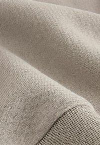 Tissu en tricot beige avec une texture douce, présentant une surface lisse et des détails de poignet côtelés. Des plis délicats créent de la profondeur dans le matériau.