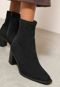 Černé semišové kotníkové boty s špičatou špičkou, blokovým podpatkem a bočním zipem. Design charakterizují minimální švy a hladká textura.