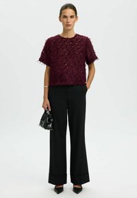 Top bordeaux dalla texture particolare con maniche corte, abbinato a pantaloni neri a gamba larga, mentre tiene in mano una piccola borsa nera.
