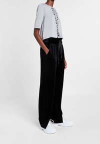 Grijze t-shirt met uitsnijdingen aan de voorkant, gecombineerd met zwarte wijde fluwelen broek met elastische tailleband en zijzakken. Witte sneakers.