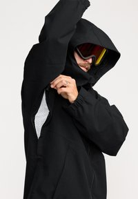 Veste noire imperméable avec une capuche ajustée, des évents zippés et des poignets élastiques. L'intérieur est doté d'une doublure en maille blanche. Modèle portant des lunettes de ski.