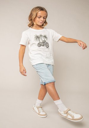 Enfant aux cheveux blonds mi-longs portant un t-shirt blanc avec un motif de palmier et de moto, un short en jean clair, des chaussettes blanches et des baskets beige.