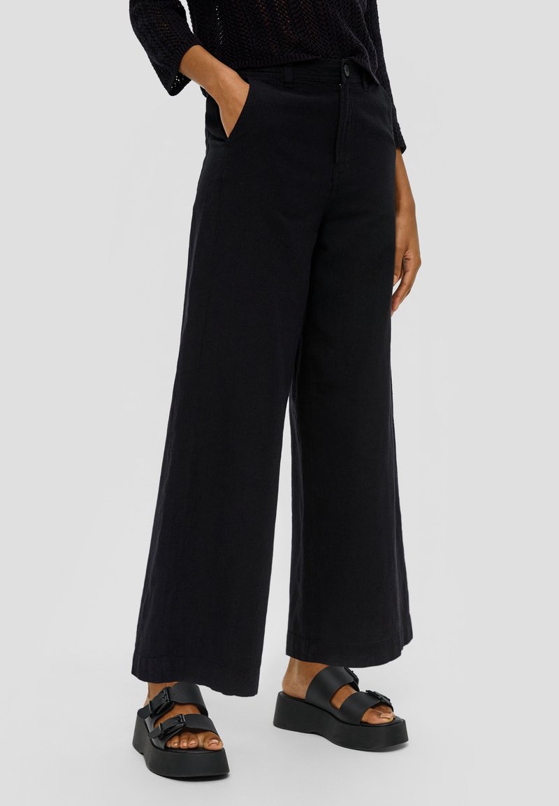 Femme portant un pantalon noir à jambes larges avec des poches et des sandales plateformes noires, debout devant un fond gris clair uni.