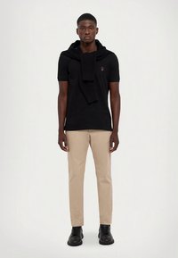 Svart kortärmad pikétröja med stickad halsduk, beige byxor och svarta skor. Har en liten logotyp på bröstet. Mjukt tyg och skräddarsydd passform.