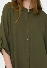 Olivgrünes Hemd zum Knöpfen mit Mandarinkragen und weitem Schnitt. Ausgestattet mit einer strukturierten Stoffqualität und runden schwarzen Knöpfen entlang der Vorderseite.