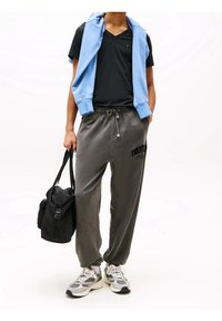 Gray jogger pants with drawstring waist and branding, paired with a black v-neck t-shirt and light blue sweatshirt draped over shoulders. Black bag and gray sneakers.