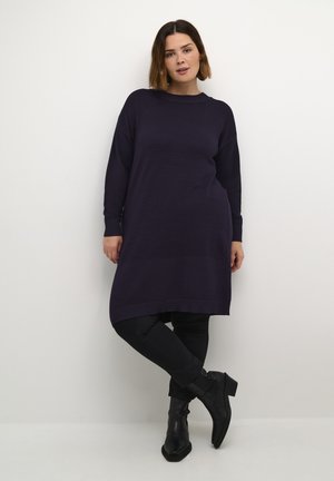 Long-sleeve tunic in dark purple ribbed fabric, featuring a relaxed fit and side slits, paired with black pants and ankle boots.
