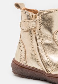 Bottine dorée métallique avec une finition en cuir texturé, zip latéral et talon de soutien. Comprend des détails perforés et une semelle en caoutchouc marron.