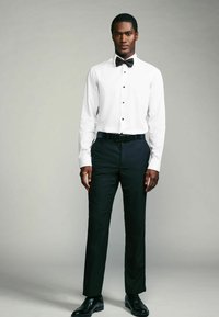 Chemise blanche avec des boutons noirs, nœud papillon noir, associée à un pantalon sombre et des chaussures noires cirées, présentant une coupe soignée et ajustée.
