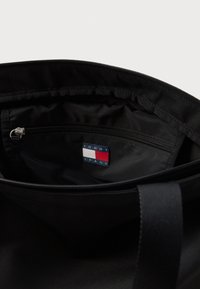 Μαύρη καμβά τσάντα εσωτερικά με φερμουάρ, που διαθέτει ετικέτα "TOMMY JEANS" σε μπλε, κόκκινο και λευκό. Λείος υφασμάτινος σχεδιασμός, ανθεκτική κατασκευή.