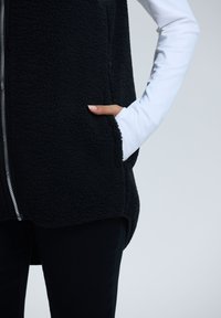 Schwarze Fleece-Weste mit Reißverschluss vorn, Seitentaschen und längerer Rückensaum, kombiniert mit einem weißen Langarmshirt darunter.