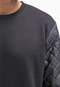 Primo piano di una persona che indossa un felpa nera con maniche in pelle trapuntata, mostrando la texture e i dettagli della zona delle spalle.