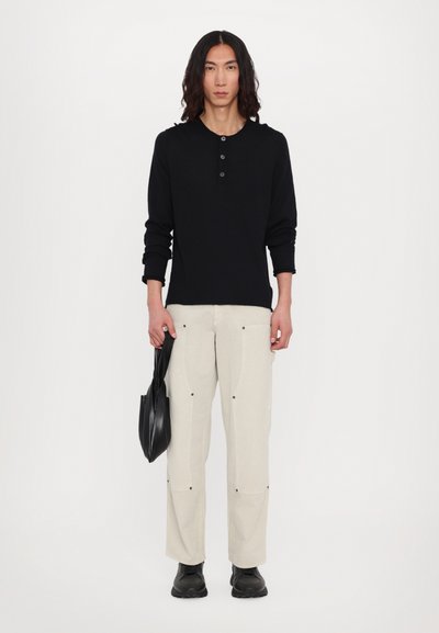 Black long-sleeve henley shirt with three buttons, beige loose-fit pants with contrasting stitching, and black boots. Holding a black bag.