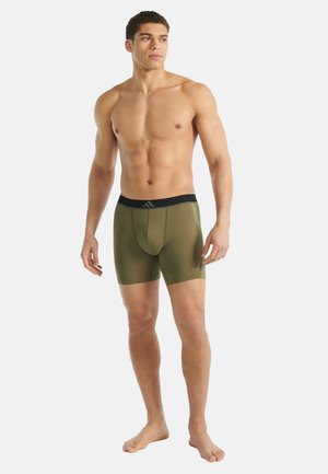 Atletische jonge man staat blootvoets in olijfgroene compressie shorts met een zwarte tailleband tegen een effen witte achtergrond.