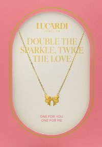 Gouden ketting met een strikvormige hanger, met een gestructureerd ontwerp. Gepresenteerd op een roze kaart met gouden tekst.