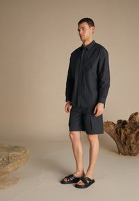 Dark, textured long-sleeve shirt, matching shorts with cargo pockets, and black sandals with thick straps. Neutral background with natural elements.