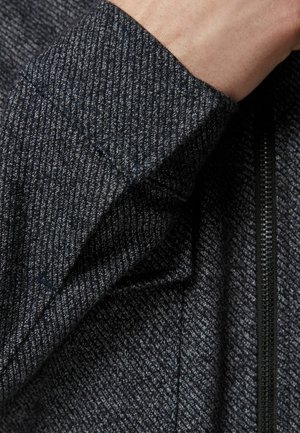 Giacca con zip grigio scuro con motivo a spina di pesce testurizzato. Presenta cuciture a contrasto e una vista ravvicinata di un polsino.