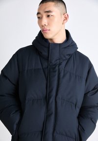 Marinblå vadderad jacka med hög krage och fastsydd huva, med intrikata quiltade sektioner och en mjuk tygtextur.