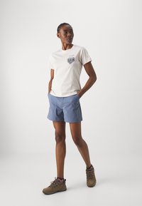 Katoenen t-shirt in off-white met grafisch detail op de borst, gecombineerd met blauwe shorts en bruine wandelschoenen.