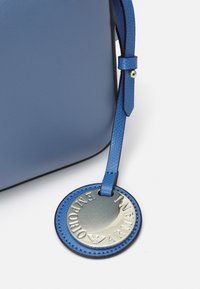 Borsa in pelle blu con un'etichetta rotonda in pelle blu coordinata e logo argento Emporio Armani appesa a una tracolla su uno sfondo bianco.