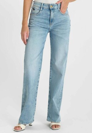 Vrouw draagt lichtblauwe spijkerbroek met een hoge taille en rechte pijpen met voorzakken, gecombineerd met witte sandalen met open tenen en hakken, staand tegen een witte achtergrond.