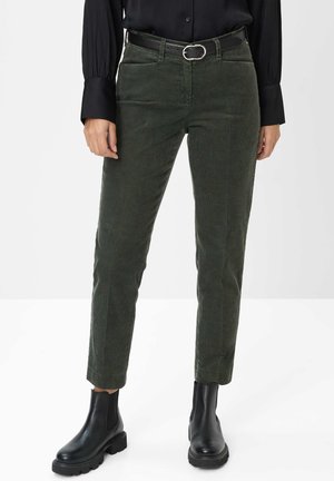 Personne portant un pantalon en velours côtelé vert foncé avec une ceinture noire, une chemise noire à manches longues et des bottines noires épaisses, debout sur un sol blanc.